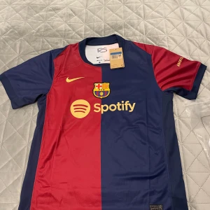 FC Barcelona hemmatröja 2023/24 - Säljer en officiell FC Barcelona hemmatröja från Nike med klassiska blå och röda vertikala fält, klubbmärke och Spotify-logga på bröstet. Kortärmad modell i Dri-FIT-material med gul Nike-logga och detaljer. Perfekt för alla Barca-fans! Oanvänd och fräsch. Köpt för 1100kr