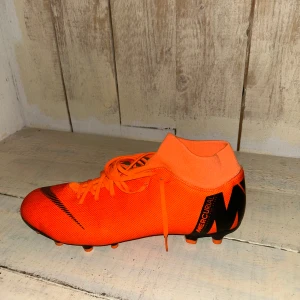 Nike Mercurial fotbollsskor  - Säljer ett par Nike Mercurial fotbollsskor i en riktigt snygg orange färg med svarta detaljer. Skorna har snörning, rund tå och platt sula med dobbar för bra grepp på planen. Perfekta för dig som vill sticka ut på fotbollsplanen.