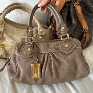 Brun Q Classic Baby Groove väska! - Snygg brun handväska i vintage skick från Marc by Marc Jacobs i äkta läder med gulddetaljer och dubbla handtag. Finns repor på lädret, se bild. Små slitningar finns på några gulddetaljer. Snygg vintage väska i en otrolig modell! Perfekt för att ge en stilren touch till din outfit. Skriv privat för fler bilder. Mått 34x18x12 (undersida). Kan hämtas hemma hos mig i Nacka eller skickas men kan tyvärr inte mötas🤍