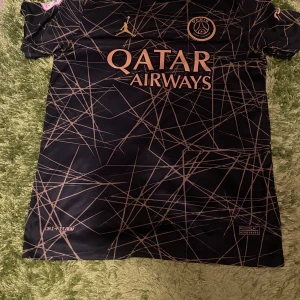 PSG fotbollströja Mbappé #7 svart/guld - Säljer en svart PSG fotbollströja med guldiga detaljer och tryck, inklusive Mbappé #7 på ryggen. Tröjan har korta ärmar, Qatar Airways sponsorlogga och ett unikt mönster i guld. Perfekt för dig som älskar fotboll och PSG.