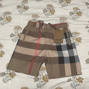 BURBERRY Shorts Helt Ny - Snygga shorts från Burberry med klassiskt rutigt mönster i beige, svart, röd och blå. Shortsen har elastisk midja med snörning och fickor både fram och bak. Perfekta för sommaren och ger en lyxig känsla till din outfit.