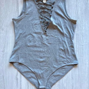 Grå bodysuit med snörning från H&M - Säljer en grå bodysuit från H&M med snygg snörning framtill i v-ringad stil. Ärmlös modell i mjuk bomullsmix, perfekt för lager-på-lager eller som statement-piece.