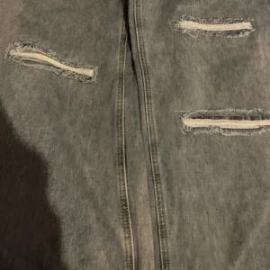 Blå slitna jeans med raka ben - Säljer ett par blåa jeans med flera slitningar på benen och klassisk femficksdesign. Jeansen har raka ben och normal passform, perfekt för en avslappnad och trendig look.