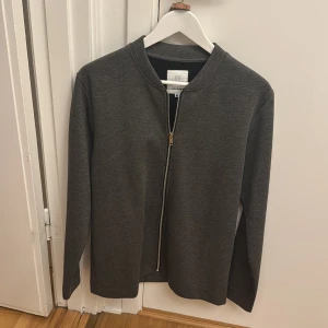 Mörkgrå zip-jacka från Elvine - Säljer en stilren mörkgrå jacka från Elvine i storlek M. Jackan har en rak passform, lång ärm och dragkedja framtill. Perfekt för dig som gillar enkel och clean stil.