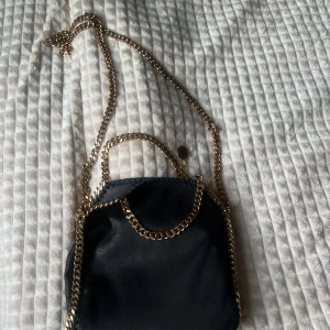 Svart axelväska från Stella McCartney - Säljer en svart axelväska från Stella McCartney med kedjedetaljer i guld och matchande axelrem. Väskan har en mjuk insida med logotypmönster och stängs med magnetknapp. Perfekt för dig som vill ha en stilren och trendig accessoar. Den är aldrig använd har köpt den på plick och den var helt ny då.