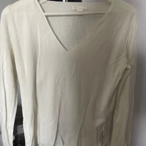 Vit v-ringad stickad tröja BASIC - Vit stickad tröja från BASIC med v-ringning och långa ärmar. Tröjan har en enkel och stilren design med små slitsar i sidorna. Perfekt för lager-på-lager och passar till många olika outfits.