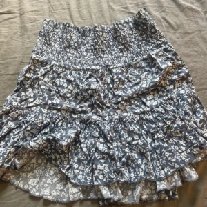 Blommig volangkjol i blått och vitt - Supersöt kort kjol med blå botten och vitt blommönster. Kjolen har bred smock i midjan och flera volanger som ger en luftig och somrig känsla. Perfekt att styla med en enkel topp eller hoodie.