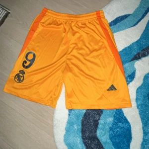 Real Madrid fotbollsshorts i orange från Adidas - Orangea fotbollsshorts från Adidas med Real Madrid-logga och nummer 9 tryckt på ena benet. Shortsen har elastisk midja och är gjorda i ett glansigt syntetmaterial. Perfekta för fotbollsträning eller match.