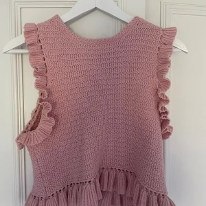 Rosa stickad klänning med volangdetaljer - Supersöt rosa stickad klänning med volanger längs ärmhål och nederkant. Perfekt för dig som gillar en romantisk och feminin stil. Klänningen har en rund halsringning och är ärmlös.