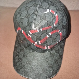 Svart keps i med orm-motiv - Snygg svart keps från med klassiskt GG-mönster och en röd och vit orm på framsidan. Kepsen har nät på sidorna och justerbar rem baktill. Perfekt för dig som vill sticka ut med en exklusiv accessoar.