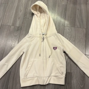 Vit velour hoodie från Juicy Couture - Säljer en vit hoodie i mjuk velour från Juicy Couture med dragkedja och huva. Framsidan har ett hjärta i strass och baksidan har ett stort strassmotiv med texten Juicy Couture. Perfekt för en trendig och avslappnad look.