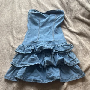 Jeansklänning med volanger  - Säljer en axelbandslös jeansklänning i ljusblå denim. Klänningen har flera lager volanger i kjolen och dragkedja bak för enkel påklädning. Perfekt för dig som gillar en cool och lekfull stil.