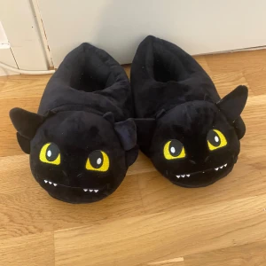 Svarta tofflor med drakmotiv - Mysiga svarta tofflor i plysch med drakansikte och gula ögon. Perfekta för att hålla fötterna varma hemma, med gulliga detaljer som öron och små tänder framtill.