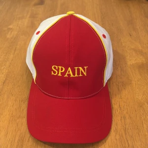 Röd och vit keps med SPANIEN-tryck - Snygg keps i rött och vitt med gul text 'SPAIN' broderat framtill. Kepsen har böjd skärm och gula detaljer längs panelerna. Perfekt för att visa ditt stöd för Spanien.