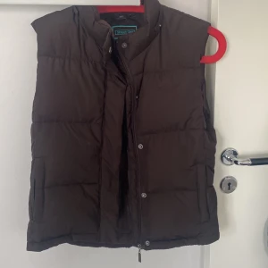 Brun dunväst från Street One - Mörkbrun dunväst från Street One med avtagbar huva . Västen har tryckknappar och praktiska fickor. 