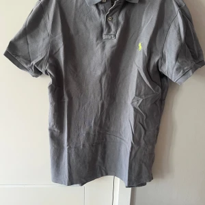 Grå pikétröja från Polo Ralph Lauren - Säljer en grå pikétröja från Polo Ralph Lauren med klassisk krage och knappslå. Tröjan har korta ärmar och en gul broderad logga på bröstet. Slim fit-modell som sitter snyggt på kroppen. Använd-med ett okej skick 