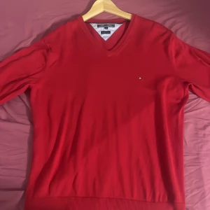 Röd v-ringad tröja från Tommy Hilfiger - Säljer en klassisk röd v-ringad tröja från Tommy Hilfiger i bomull. Tröjan har lång ärm och en liten broderad logga på bröstet. Perfekt för dig som gillar stilrena och tidlösa plagg.