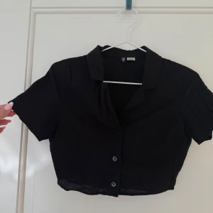 Svart croppad skjorta från H&M Divided - Svart croppad skjorta från H&M Divided med korta ärmar och klassisk krage. Skjortan har knäppning framtill och är tillverkad i en blandning av bomull och linne, vilket gör den luftig och skön. Perfekt för en trendig och avslappnad look.