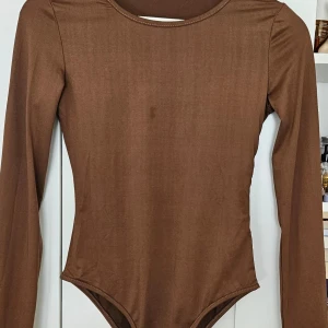 Brun långärmad bodysuit - Säljer en stilren brun långärmad bodysuit med rund halsringning. Perfekt att styla med jeans eller kjol för en trendig look. Mjuk och stretchig kvalitet som sitter snyggt på kroppen.