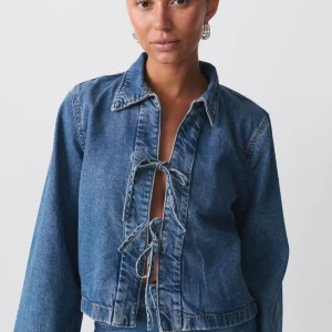 Blå croppad jeansjacka med knyt - Trendig blå jeansjacka med croppad passform och knyt framtill istället för dragkedja eller knappar. Jackan har klassisk krage och långa ärmar. Perfekt för dig som vill ha en unik denimlook.