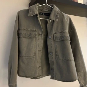 Grön/grå jeansjacka från Zara - Säljer en grön/grå jeansjacka från Zara med två stora bröstfickor och snygga broderade detaljer på ficklocken och kragen. Jackan har klassisk krage, knäppning framtill och raka ärmar. Den är för kort för mig så därför säljer jag den