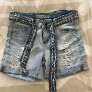 Ljusblå jeansshorts med mönstrat bälte från Amisu - Säljer ett par ljusblå jeansshorts från Amisu med slitna detaljer och upprullade benslut. Shortsen har ett blåvitt mönstrat tygbälte och klassiska fem fickor. Perfekta för sommaren!