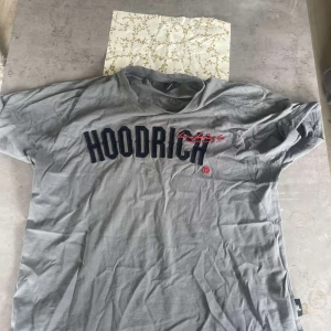 Grå t-shirt från Hoodrich - Säljer en grå t-shirt från Hoodrich med svart och röd logga framtill och tryck på ärmen. T-shirten har normal passform och är tillverkad i mjuk bomull. Perfekt för en avslappnad streetwear-look.