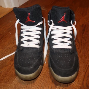 Svarta Nike Air Max sneakers med röd detalj - Säljer ett par svarta Nike Air Max sneakers med vita snören och röda detaljer, inklusive den klassiska Jumpman-loggan på plösen. Skorna har högt skaft och en robust sula med grå inslag. Perfekta för dig som gillar sportig stil.