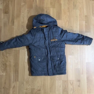 Grå vinterjacka från LC Waikiki - Mörkgrå vinterjacka från LC Waikiki med huva och orange fleecefoder. Jackan har två stora fickor framtill med knappar och dragkedja. Märke på ärmen och broderad text på bröstet. Perfekt för kalla dagar.