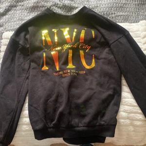 Svart sweatshirt med guldtext NYC - Svart sweatshirt med stora glansiga guldiga bokstäver 'NYC' och texten 'New York City' på ryggen. Tröjan har rund hals och långa ärmar, perfekt för en cool och avslappnad stil.