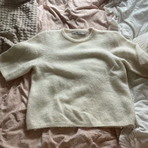 Vit stickad tröja med kort ärm - Mysig vit stickad tröja med rund halsringning och korta ärmar. Tröjan har en rak passform och är tillverkad i ett fluffigt, mjukt material som känns lyxigt. Perfekt för lager-på-lager och enkel att styla till olika outfits.