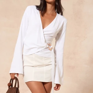 Vit omlottopp med knytning - Säljer en stilren vit omlottopp med lång ärm och v-ringning. Toppen har knytning i sidan med dekorativ detalj och är tillverkad i ett mjukt tyg.aldrig använd lappen är kvar! 