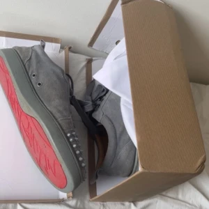 Graå loubitons - Säljer ett par gråa sneakers i mocka med coola nitar framtill och klassisk röd sula. Skorna har snörning och rund tå, vilket ger en unik och edgy look. Perfekta för dig som vill sticka ut med din stil.