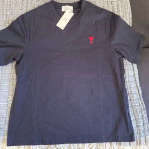 Mörkblå t-shirt från Ami Paris med rött hjärta - Säljer en mörkblå t-shirt från Ami Paris med broderad röd hjärtdetalj och bokstaven A på bröstet. Klassisk passform och rund halsringning. Perfekt för en stilren och trendig look.