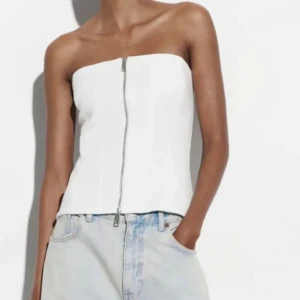 Vit tubtopp med dragkedja framtill - Säljer en snygg vit tubtopp med dragkedja framtill. Toppen är axelbandslös och har en stilren, figurnära passform som passar perfekt till både jeans och kjol. Enkel att matcha och ger en trendig look.