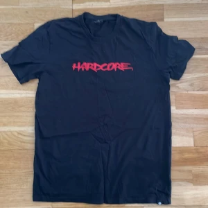 Svart t-shirt med röd text från Stanley/Stella - Säljer en svart t-shirt från Stanley/Stella i ekologisk bomull med trycket 'HARDCORE' i rött framtill. Klassisk passform och rund halsringning. 