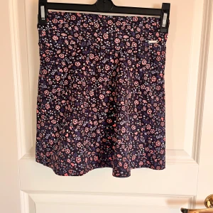 Blommig kortkjol från aimn - Supersöt kortkjol från aimn med mörkblå botten och små blommor i rosa, rött och vitt. Kjolen har en lätt utställd form och är tillverkad i ett mjukt, stretchigt material. Perfekt för dig som gillar sportig stil med en feminin touch.