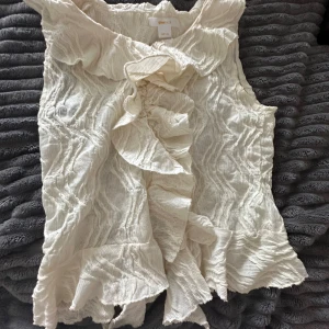 Vit volangtopp från Gina Tricot - Säljer en vit ärmlös topp från Gina Tricot med volangdetaljer längs krage och framtill. Toppen har en strukturerad, lätt skrynklig yta och är perfekt för dig som gillar romantiska detaljer.