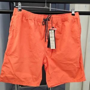 Orange shorts från O'Neill med snörning - Säljer ett par fräscha orange shorts från O'Neill med elastisk midja och svart snörning. Shortsen har sidofickor och bakfickor, perfekta för sommardagar. Lätta och bekväma, passar till strand eller stad. Vintage effekt. Bomull material.
