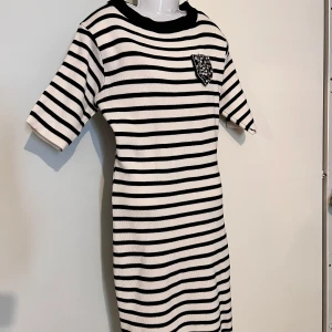 Randig ribbad klänning med pärldetalj - Säljer en svartvit ribbad klänning med horisontella ränder och en dekorerad pärlficka på bröstet. Klänningen har korta ärmar och rund halsringning. Perfekt för dig som gillar klassisk stil med en twist.