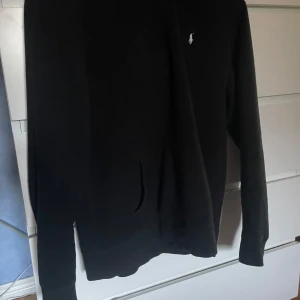 Svart hoodie från Ralph Lauren - Svart hoodie från Ralph Lauren med klassisk vit logga på bröstet. Tröjan har huva och långa ärmar samt en stor ficka framtill. Perfekt för en avslappnad stil. Pris ej hugget i sten!!