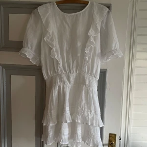 Vit volangklänning med kort ärm - Superfin vit klänning med volangdetaljer och korta ärmar. Klänningen har smock i midjan, rund halsringning och knytning i nacken. Den är lätt och luftig, perfekt för varma dagar eller sommarens tillfällen. Knappt använd och i gott skick. 💗