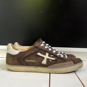 Bruna sneakers från Premiata - Säljer ett par bruna sneakers från Premiata i mocka med vita detaljer och snörning. Skorna har en beige insida och vit, mönstrad hälkappa med sprucken effekt. Klassisk rund tå och platt sula. Perfekta för dig som gillar stilrena sneakers. Köpte dem med gråa skosnören, men bytte ut dem för vita. Men självklart får ni med original skosnören också. För fler bilder, frågor eller prisförslag är det bara att skriva! 😁Priset är inte fast.