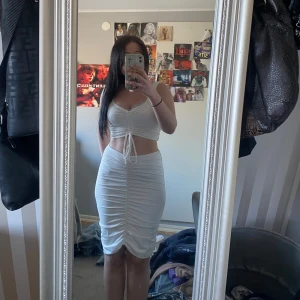 Vit omlottklänning med knyt och rynkade detaljer - Säljer en vit två delad klänning med omlott-topp, knyt framtill och rynkade detaljer längs både överdel och kjol. Klänningen är ärmlös och har en tight passform som framhäver figuren. Perfekt för sommarens tillfällen!