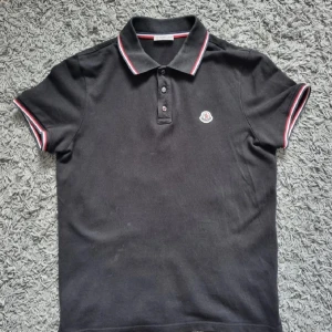 Moncler piké  - Säljer en svart pikétröja från Moncler med klassisk krage och tre knappar framtill. Tröjan har Moncler-logga på bröstet och röda samt vita detaljer på krage och ärmslut. Perfekt för en stilren och sportig look.