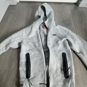 Grå nike tech fleece  - Säljer en grå hoodie från Nike med svarta dragkedjor på fickorna och Nike-logga på bröstet. Jackan har huva och långa ärmar, perfekt för en sportig stil. Tillverkad i mjukt material för extra komfort. Är använd ett par gånger. Pris kan diskuteras