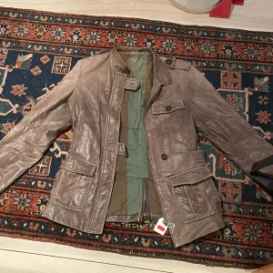 Brun skinnjacka  - Säljer en brun skinnjacka i äkta läder med flera fickor och snygga knapplås framtill. Jackan har hög krage, lång ärm och markerade sömmar. Originellt ifrån RocknBlue. Jackan är i mycket gott skick förutom att en söm vid ena framfickan har gått upp lite grann. (Se sista bilden)