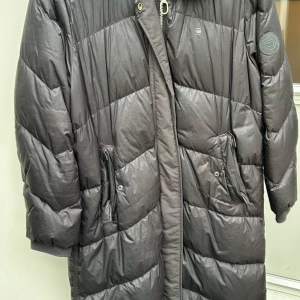 Svart lång dunjacka från G-Star RAW - Säljer en svart lång dunjacka från G-Star RAW med dragkedja och tryckknappar. Jackan har två stora fickor framtill och en hög krage. Perfekt för kalla dagar och riktigt skön att bära.