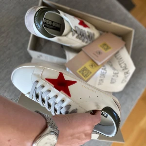 Vita sneakers med röd stjärna från Golden Goose - Säljer ett par vita sneakers från Golden Goose med en röd stjärna på sidan, grå snörning och mörkgrön häl. Skorna har en platt sula i gummi och klassisk rund tå. Perfekta för dig som gillar en sportig och trendig look.