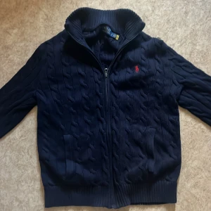 Mörkblå stickad kofta från Polo Ralph Lauren - Säljer en svinsnygg mörkblå stickad kofta från Polo Ralph Lauren med dragkedja och krage. Tröjan har kabelstickat mönster, två fickor framtill och en liten röd broderad logga på vänster bröstdel. Riktigt stilren tröja som är i storlek S. Skriv vid frågor!😊